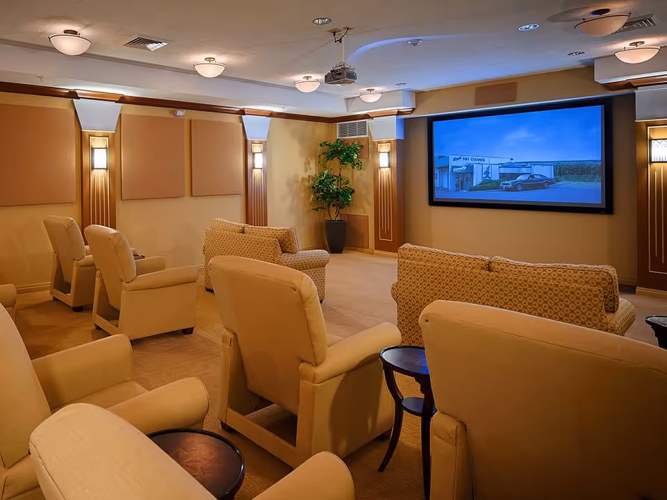 A cozy home theater room with multiple beige armchairs and patterned sofas arranged facing a large screen on the wall. The room has warm lighting with wall sconces and ceiling lights, a potted plant in the corner, and a ceiling-mounted projector.