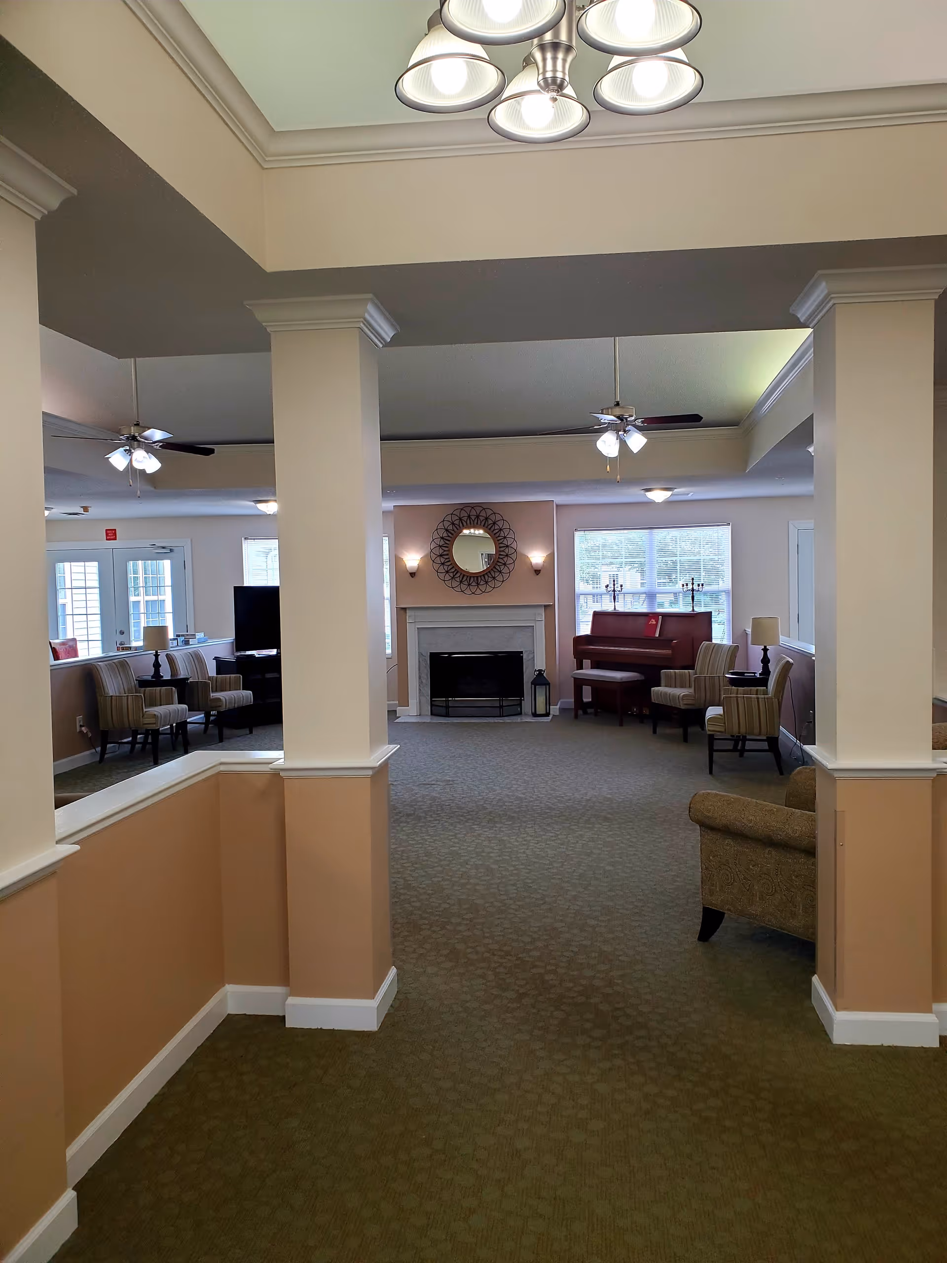 Interior view of a senior living facility common area with beige walls and carpeted floor. The room features a fireplace with a decorative round mirror above it, several armchairs, a piano near the window, ceiling fans with lights, and a chandelier hanging from the ceiling.