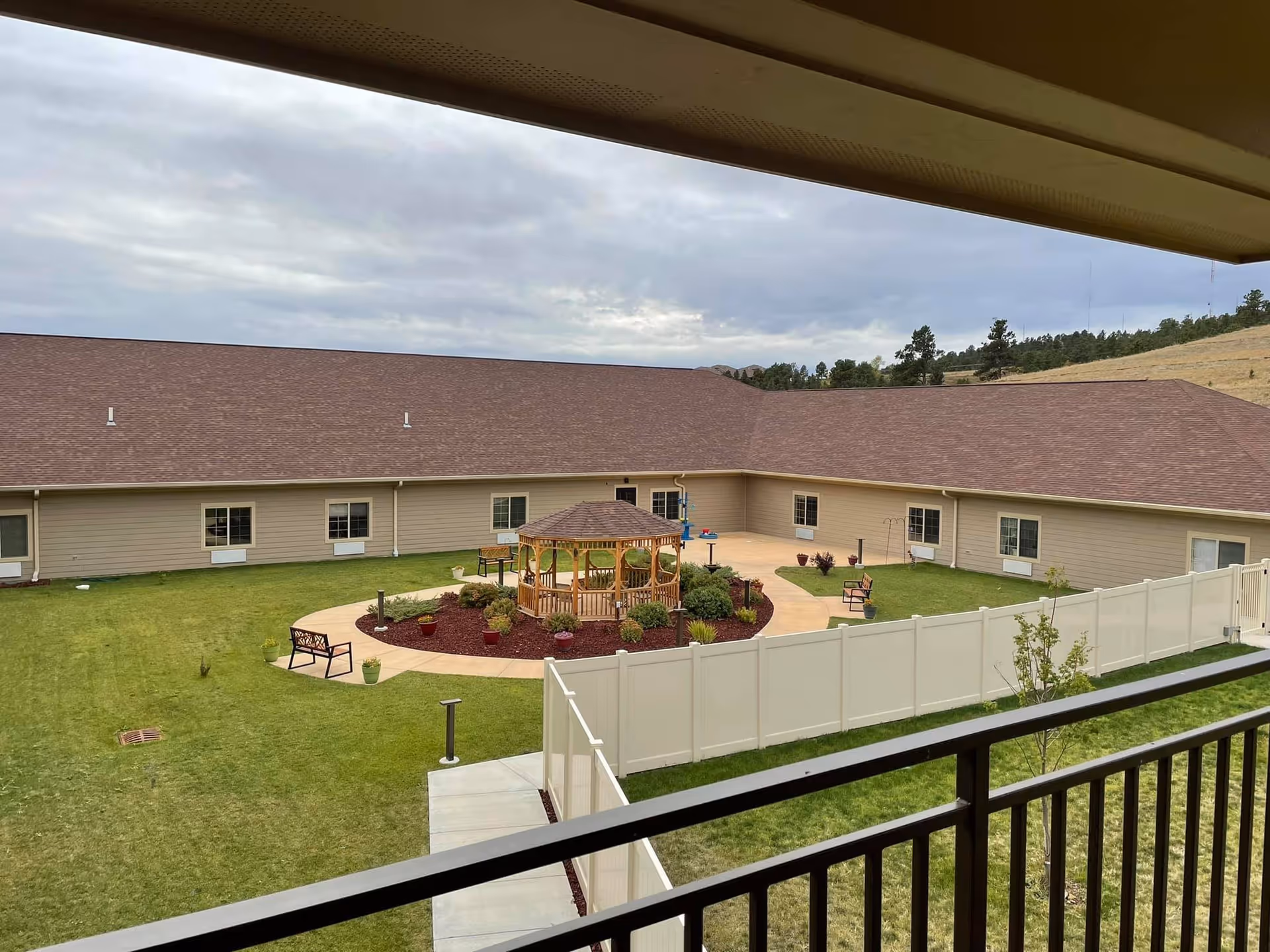 View of a courtyard at The Village at Skyline Pines Assisted Living & Memory Care, featuring a central wooden gazebo surrounded by a circular walkway, benches, potted plants, and green grass. The courtyard is enclosed by a beige building with multiple windows and a brown roof, with a white fence and railing in the foreground.
