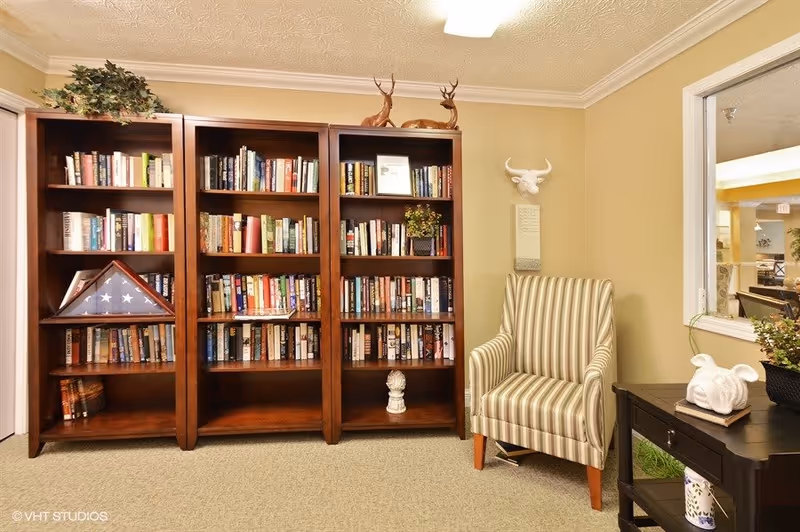 A cozy reading nook featuring three wooden bookshelves filled with books, a folded American flag in a display case, and decorative items including two deer figurines on top of the shelves. To the right is a striped armchair next to a small black side table with a white rabbit sculpture and a potted plant. A window on the right wall looks into another room.