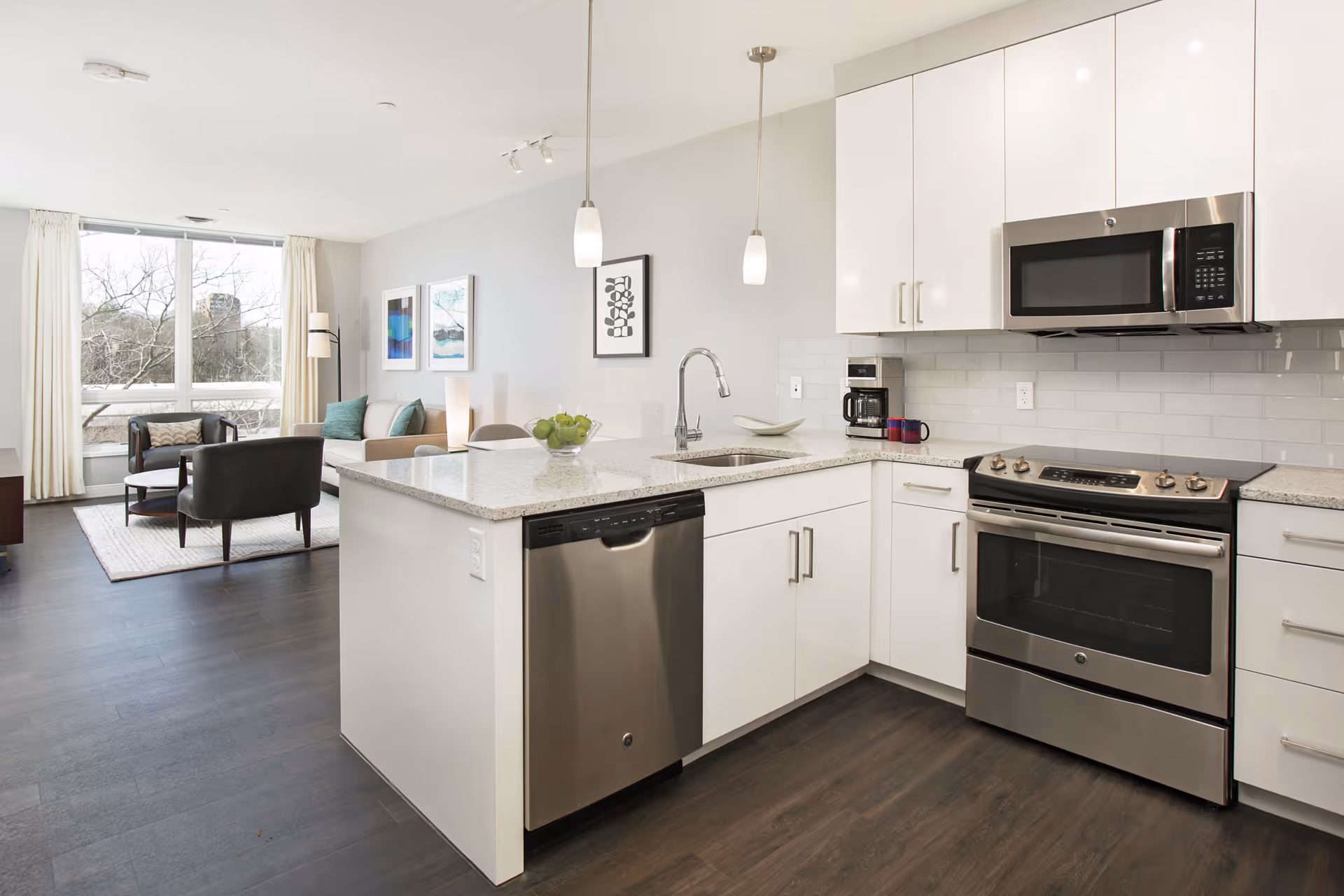 Modern open-concept kitchen and living room area with white cabinets, stainless steel appliances including a dishwasher, oven, and microwave. The kitchen island has a sink and two pendant lights hanging above. The living room features a large window with white curtains, a beige sofa, two black chairs, a floor lamp, and framed artwork on the wall.