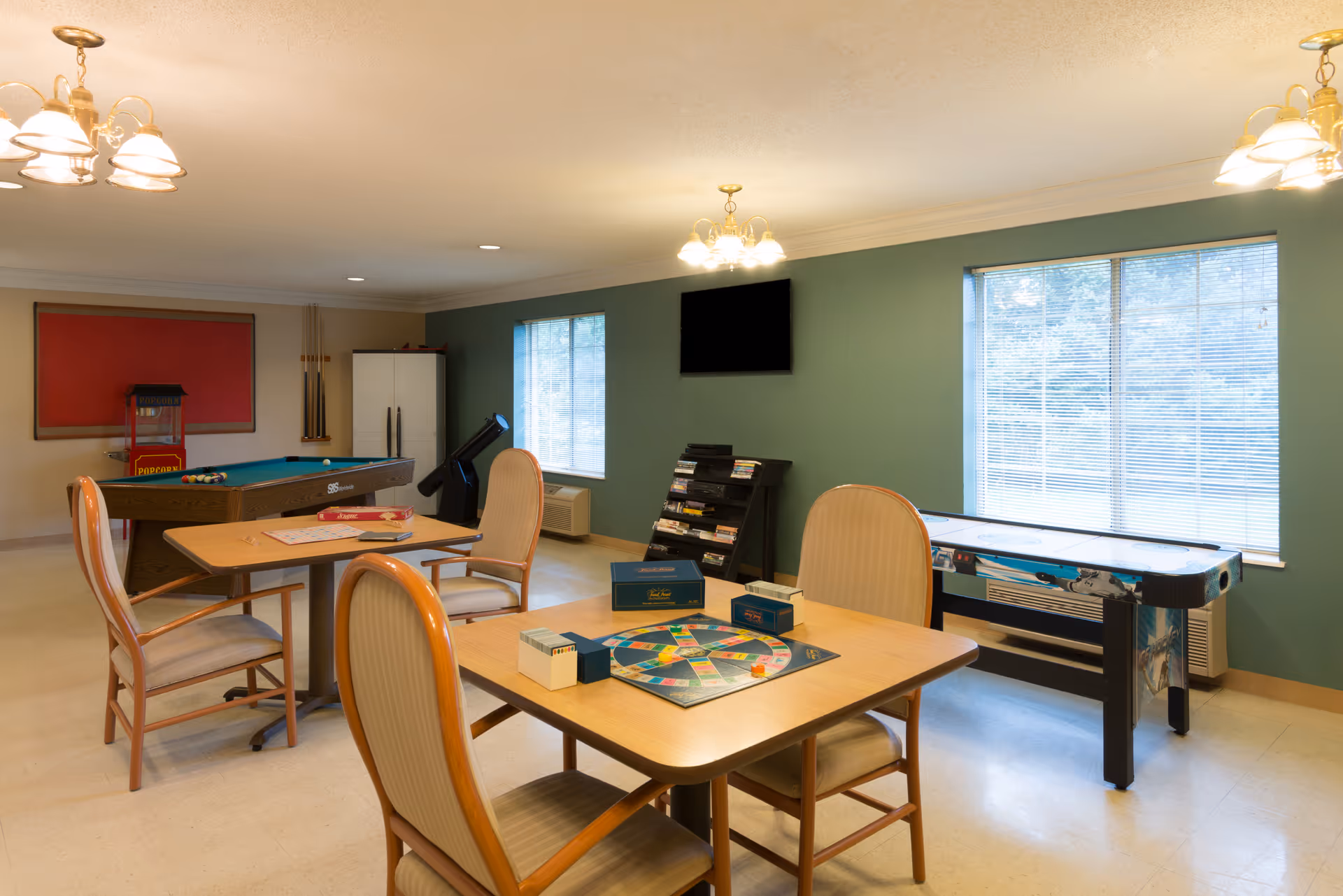 A recreational room with two tables and chairs, one table has a board game set up. The room also features a pool table, a popcorn machine, a shuffleboard table, a wall-mounted TV, and a rack with DVDs or games. The walls are painted green and beige, and there are two large windows letting in natural light.