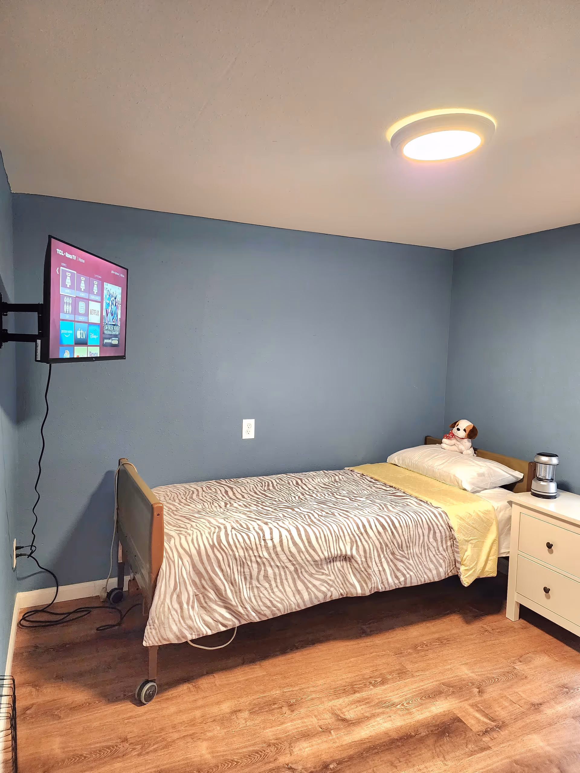 A small bedroom with blue walls and wooden floor. There is a single bed with a zebra-patterned blanket, a yellow sheet, and a pillow with a stuffed dog toy on it. A white nightstand with two drawers and a small lamp is next to the bed. A flat-screen TV is mounted on the wall opposite the bed. The room is lit by a round ceiling light.
