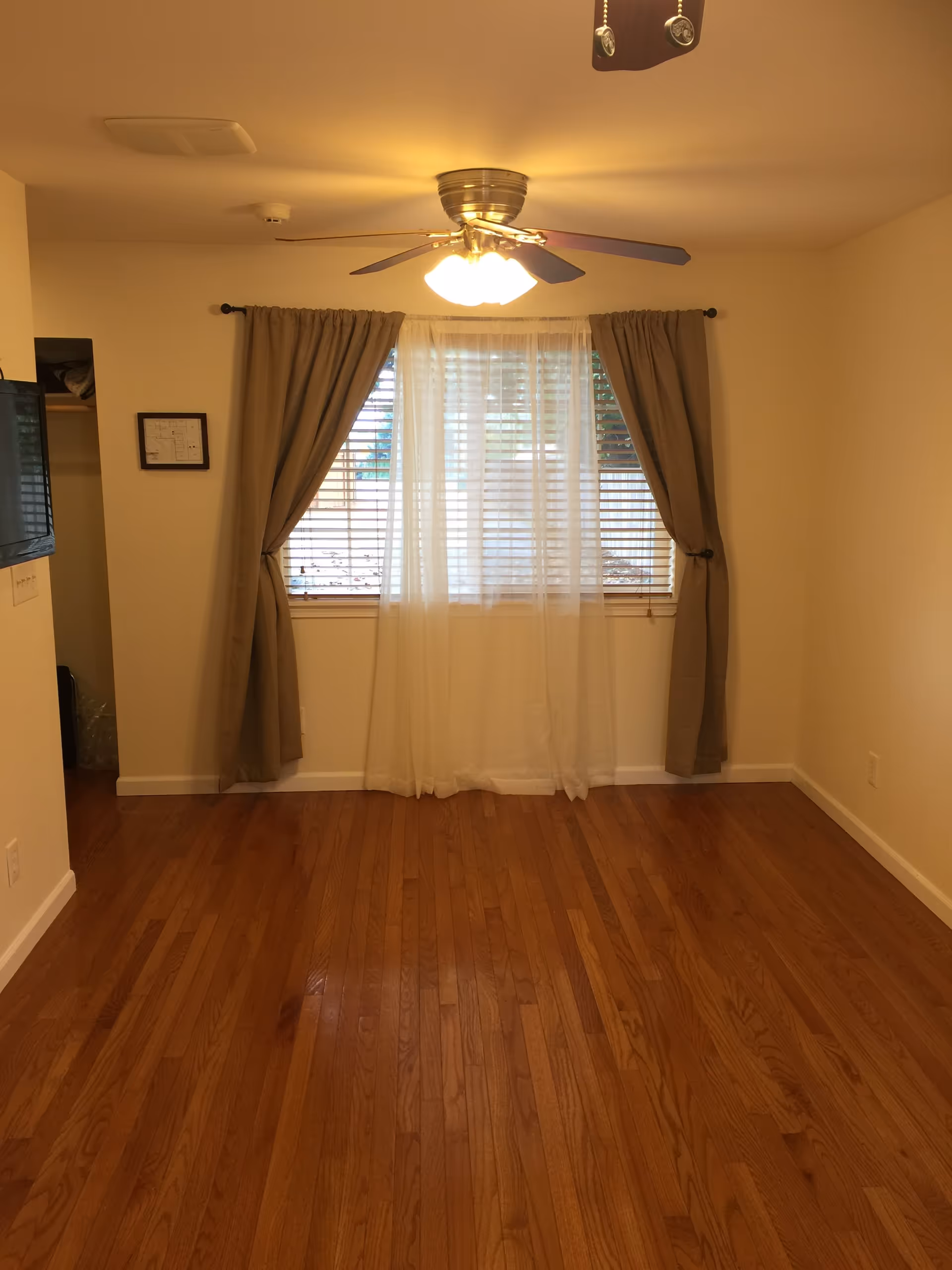 Empty room with hardwood floors, a window with blinds and curtains, and a ceiling fan overhead.