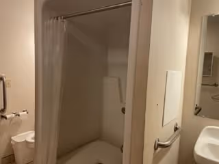 A small bathroom with a shower area featuring a white curtain, a wall-mounted soap dish, and a grab bar. There is a sink with a mirror above it and a hand towel holder on the wall.
