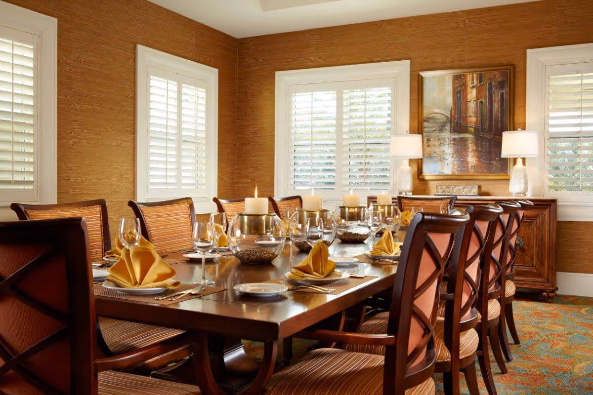 A formal dining room with a long wooden table set for a meal. The table is adorned with yellow folded napkins, wine glasses, plates, and candles in glass holders. The room has warm brown walls, three windows with white shutters, two table lamps on a sideboard, and a framed painting on the wall.