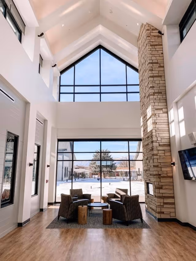 A bright and spacious common area with high vaulted ceilings and large windows letting in natural light. The room features a stone fireplace on the right side, four cushioned armchairs arranged around a small round table on a rug, and wooden flooring. Outside the windows, a snowy landscape with trees and buildings is visible.