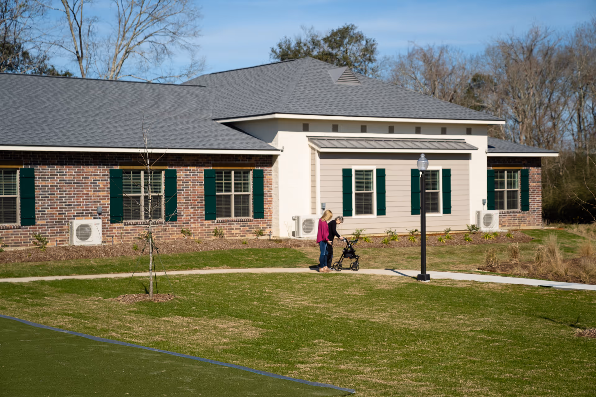 Two women walking on a paved pathway outside a single-story brick and siding building with green window shutters. One woman is using a walker while the other is assisting her. The area has a grassy lawn, a young tree, and a lamp post, with leafless trees in the background under a clear blue sky.