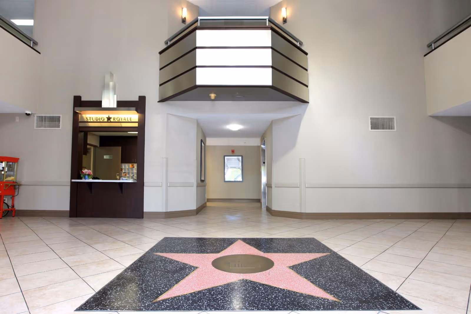 Interior lobby area of Studio Royale facility featuring a tiled floor with a large pink star design in the center. There is a reception window with a sign above it that reads 'Studio Royale' on the left side, and a hallway leading further into the building. The walls are light-colored with minimal decor, and there are two balconies visible on the upper level.