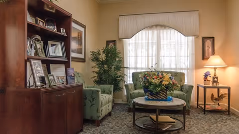 A cozy living room area with two green upholstered armchairs and a matching loveseat arranged around a round coffee table with a floral centerpiece. A wooden cabinet with framed photos and awards is on the left, and a side table with a lamp and decorative items is on the right. A large window with sheer curtains and a valance lets in natural light, and a potted plant adds greenery to the space.