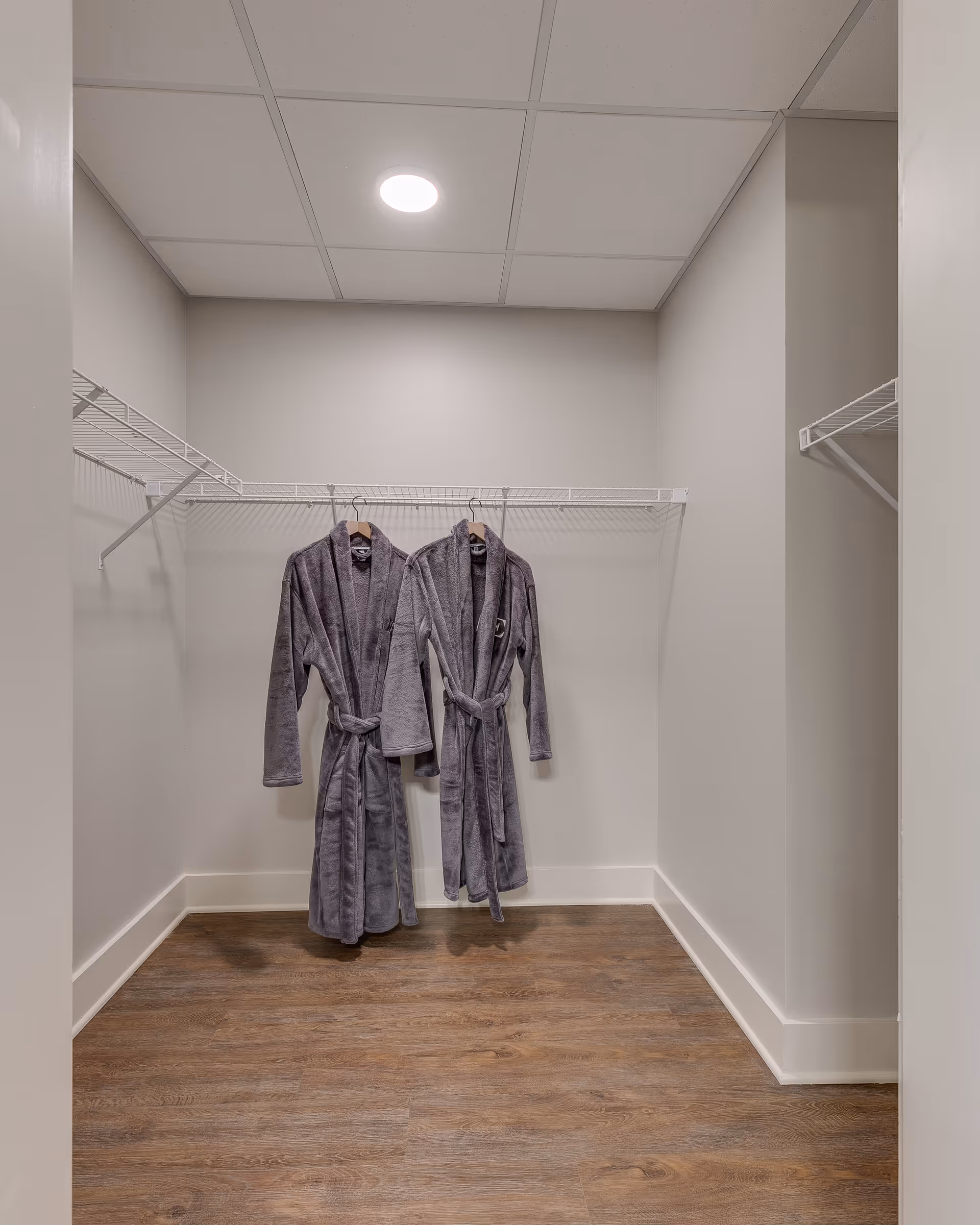 A small walk-in closet with light gray walls, wood flooring, and white wire shelving on three sides. Two gray bathrobes are hanging on the middle shelf against the back wall under a ceiling light.