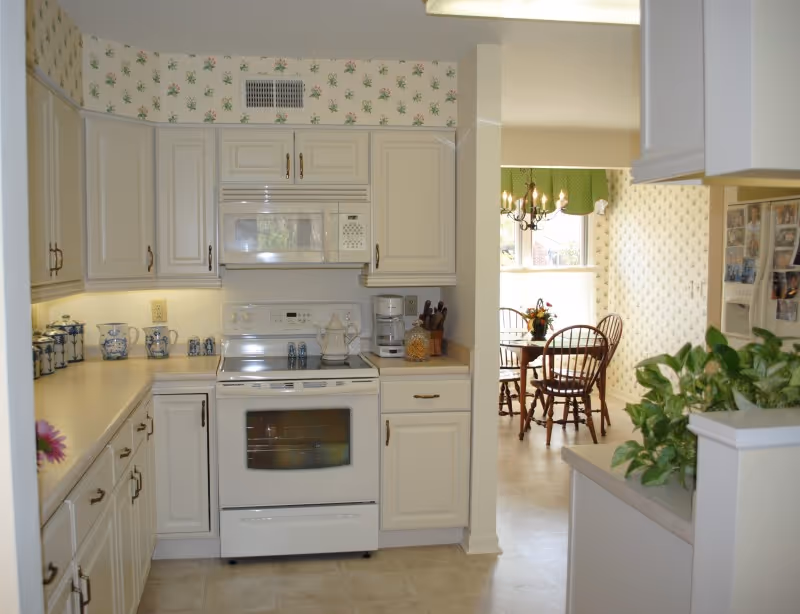 A bright kitchen with white cabinets and appliances including a stove and microwave. The countertops have decorative blue and white ceramic containers and a coffee maker. To the right, there is a small dining area with a wooden table and chairs near a window with green valance curtains. A green plant is visible on the right side of the image.
