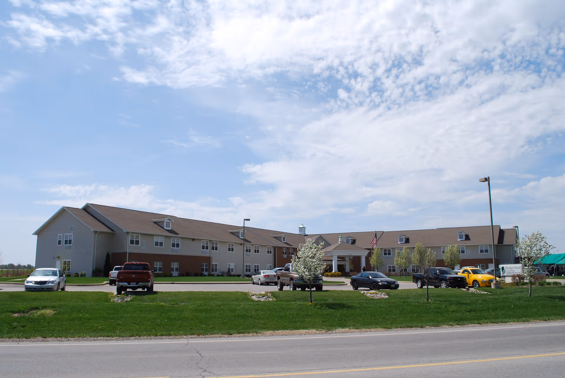 Exterior view of a large two-story senior living facility building with a brown roof and beige siding, surrounded by a green lawn and several small trees. Multiple cars are parked in front of the building under a partly cloudy sky.
