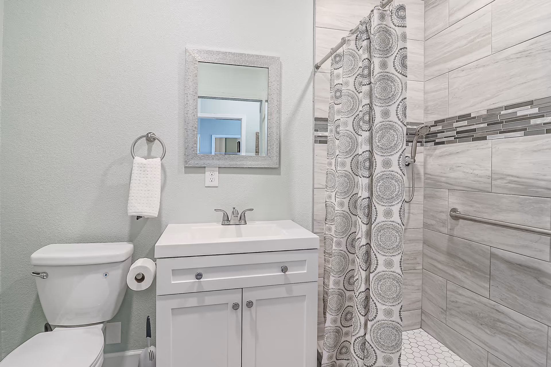 A clean and modern bathroom featuring a white toilet, a white vanity with a sink and silver faucet, a rectangular mirror with a textured frame above the sink, a towel ring with a white towel, and a shower area with a patterned shower curtain and tiled walls with a horizontal mosaic accent strip.