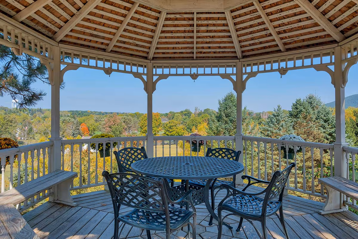 Wooden gazebo with a round metal table and chairs overlooking a tree-filled landscape.
