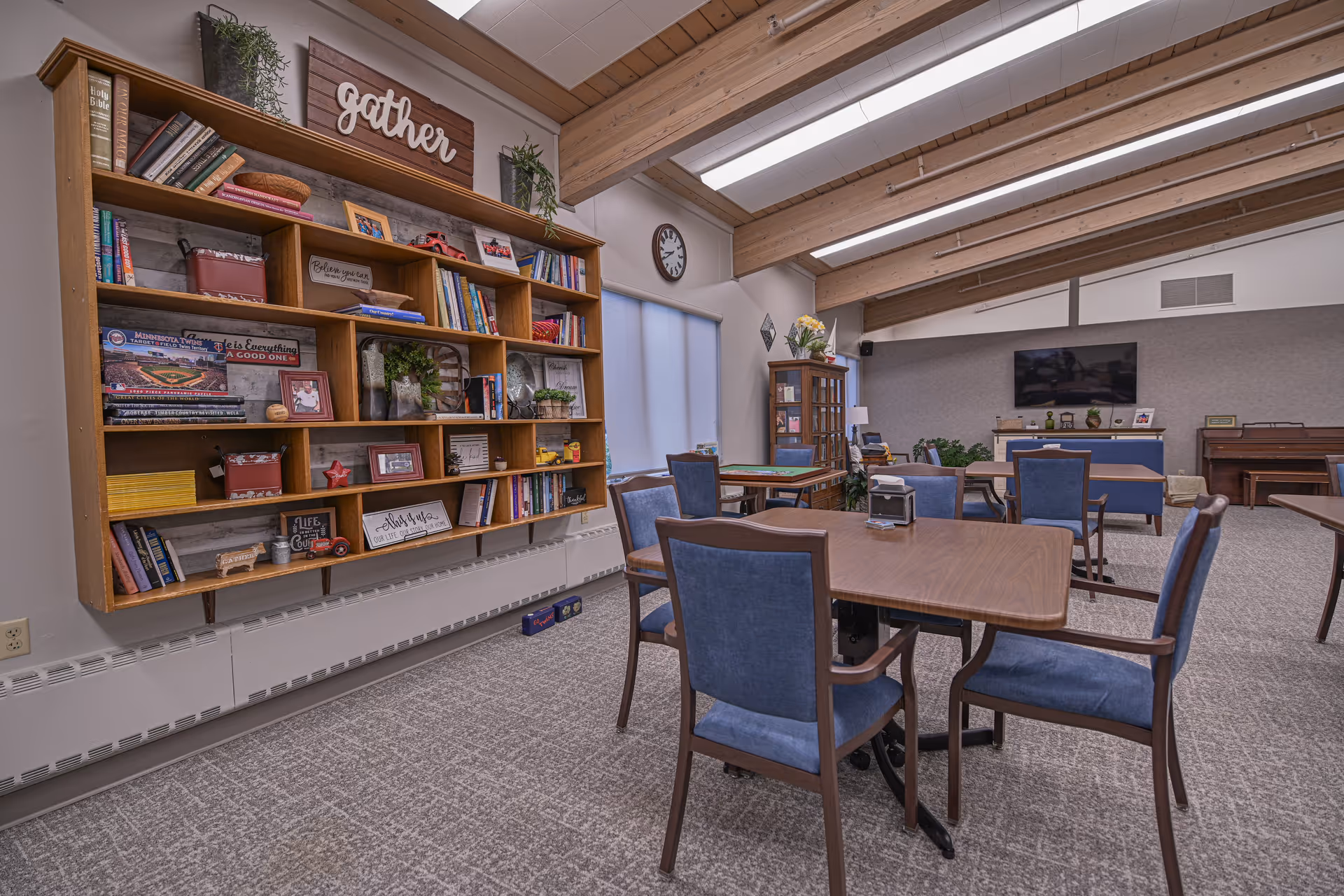 A cozy common area with wooden tables and blue cushioned chairs arranged for seating. A large wooden bookshelf filled with books, decorative items, and plants is mounted on the wall with a sign that says 'gather' above it. The room has a high ceiling with exposed wooden beams and skylights. A clock is mounted on the wall near a window with blinds. In the background, there is a TV mounted on the wall, a piano, and additional seating.