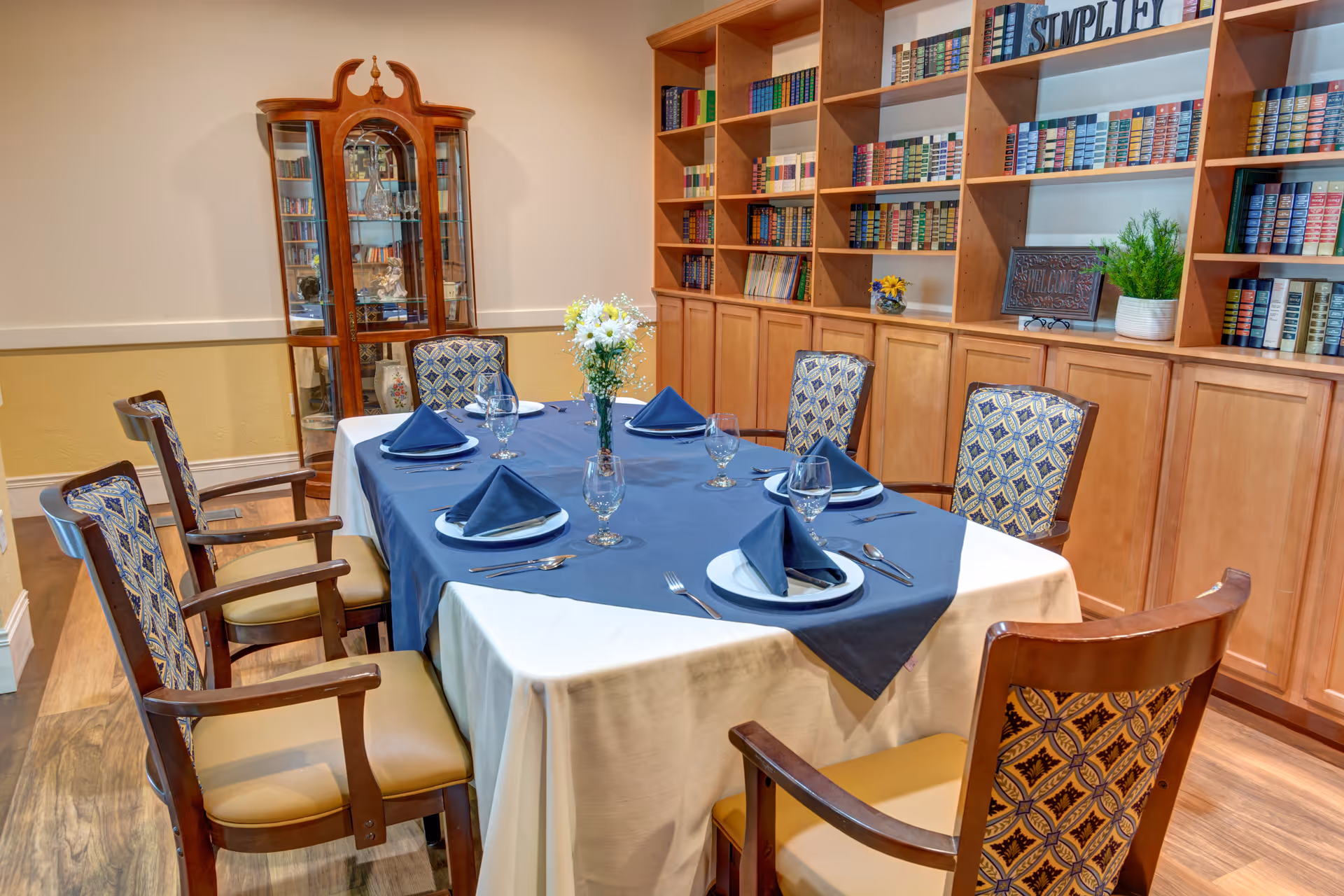 A dining room with a rectangular table set for six people. The table is covered with a white tablecloth and a blue table runner, with blue folded napkins, plates, glasses, and silverware arranged neatly. There are six wooden chairs with patterned upholstery around the table. In the background, there is a wooden cabinet filled with books and decorative items, and a glass display cabinet in the corner.