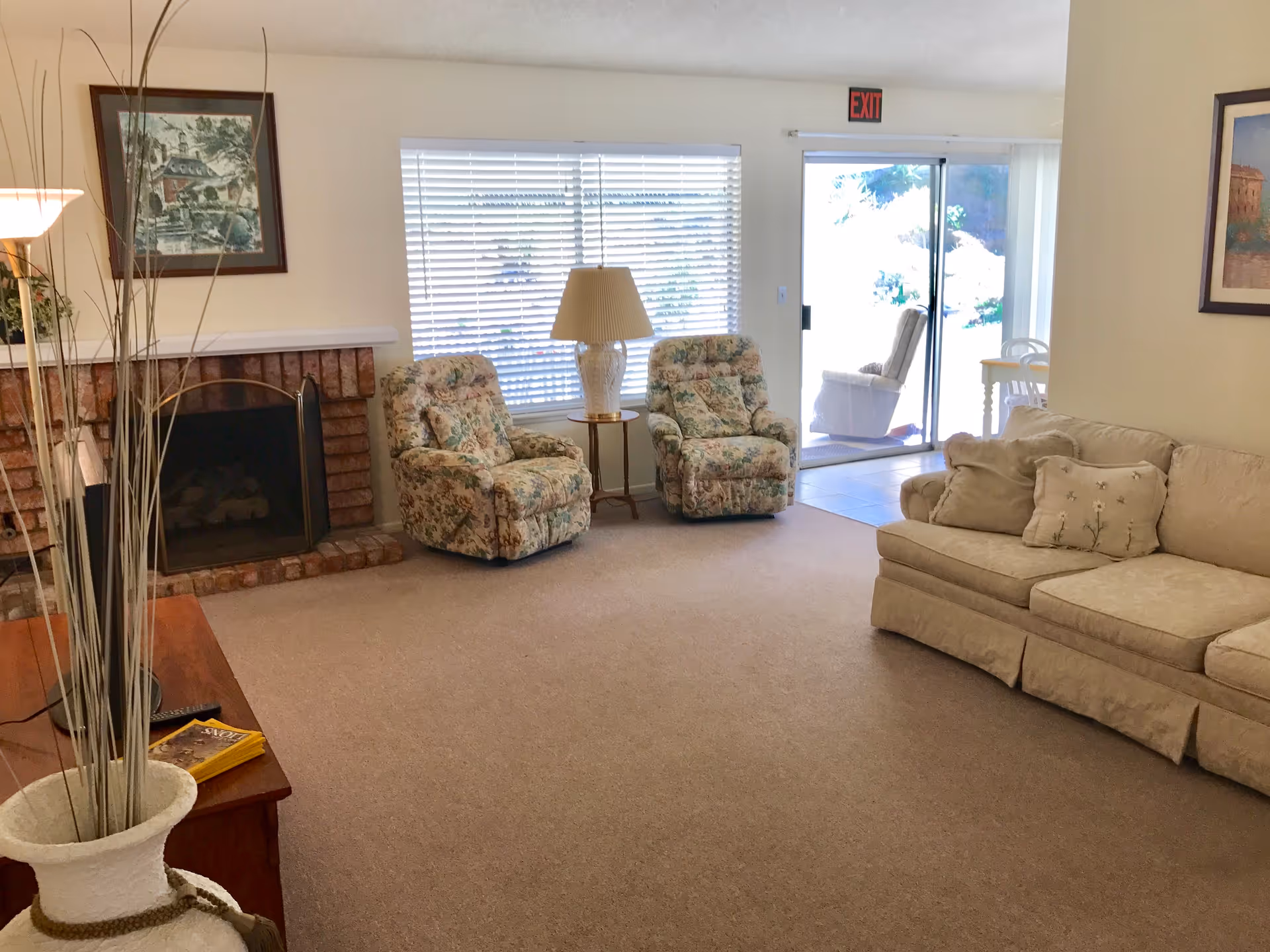 A cozy living room with beige carpet, a beige sofa with decorative pillows, two floral patterned armchairs, a wooden side table with a lamp, a brick fireplace with a framed picture above it, a large window with blinds, and a sliding glass door leading to an outdoor area.