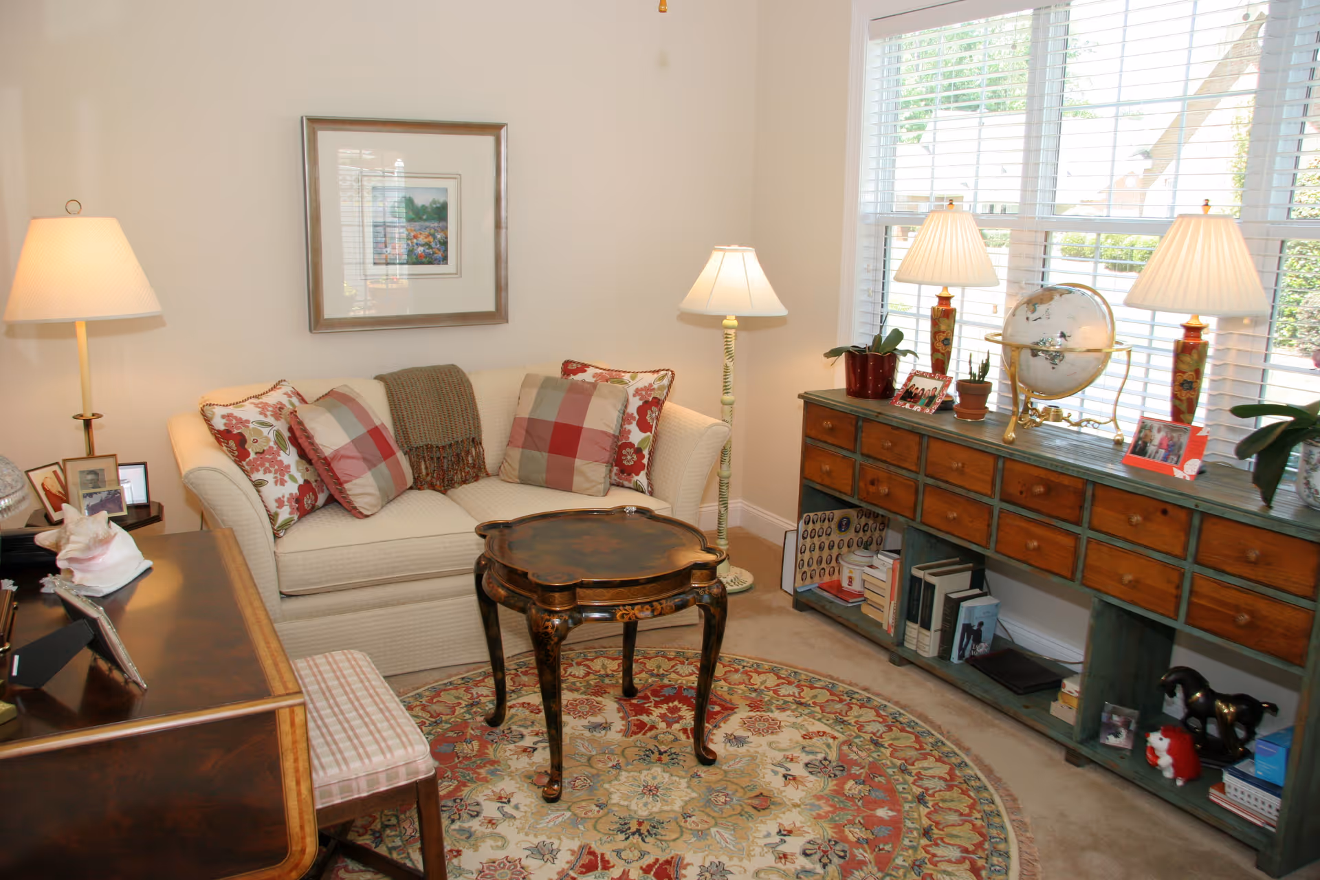 A cozy living room with a cream-colored sofa adorned with patterned pillows and a throw blanket. In front of the sofa is an ornate wooden coffee table on a round floral rug. To the right, a large window with white blinds lets in natural light, illuminating a long wooden console table with multiple drawers, decorated with table lamps, plants, a globe, and framed photos. A floor lamp stands next to the sofa, and a wooden desk with a chair is partially visible on the left side.
