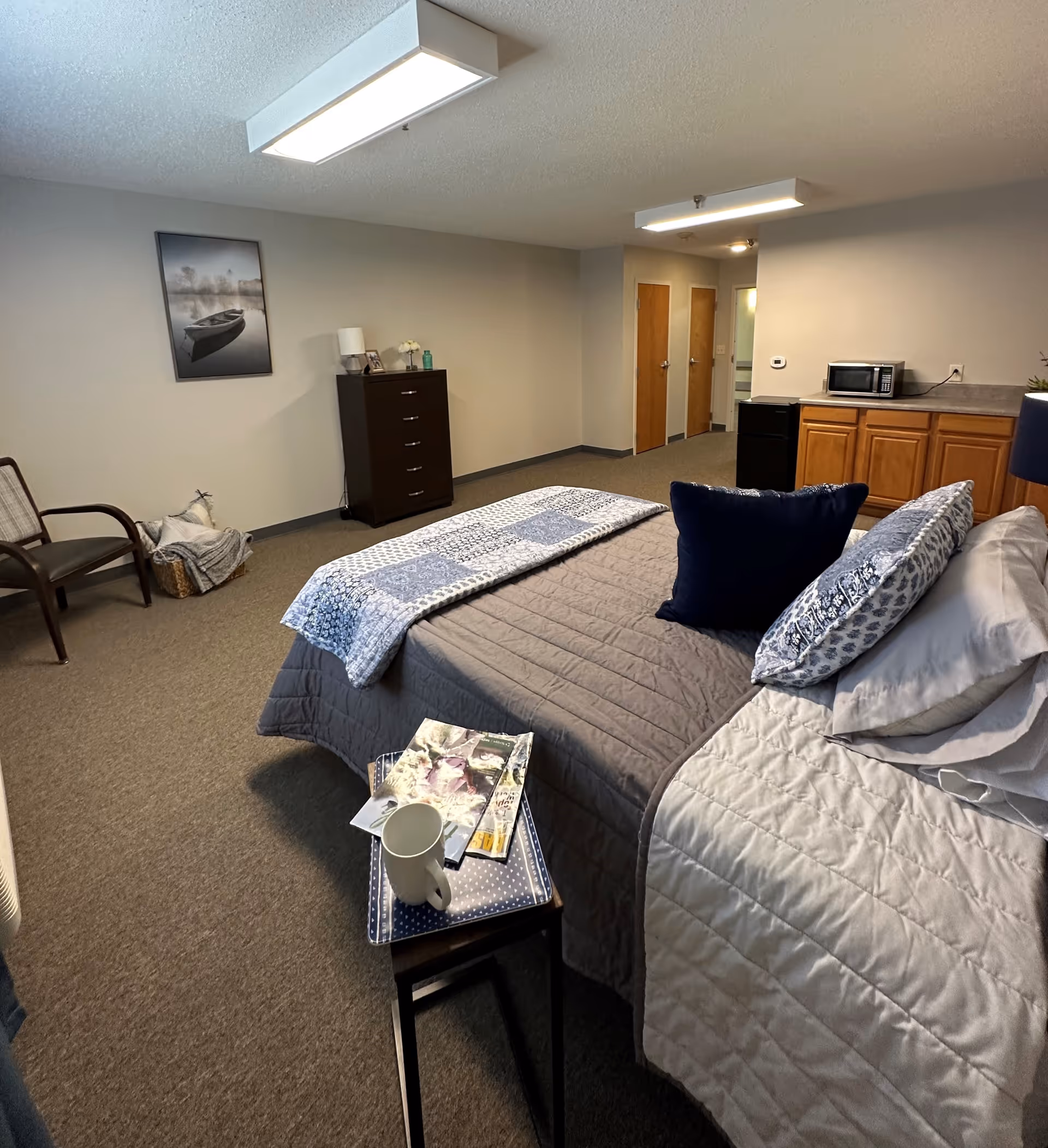 A senior living facility bedroom with a neatly made bed featuring gray and blue bedding and pillows. A small side table next to the bed holds a white mug and some magazines. In the background, there is a dark wooden dresser with a lamp and decorative items on top, a chair with a blanket and pillow, and a kitchenette area with wooden cabinets, a microwave, and a mini fridge. The walls are light gray, and there is a framed picture of a boat on the wall.