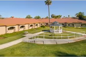 Sunlit courtyard featuring a white gazebo, green lawns, walkways, and single-story buildings with red-tiled roofs and palm trees.