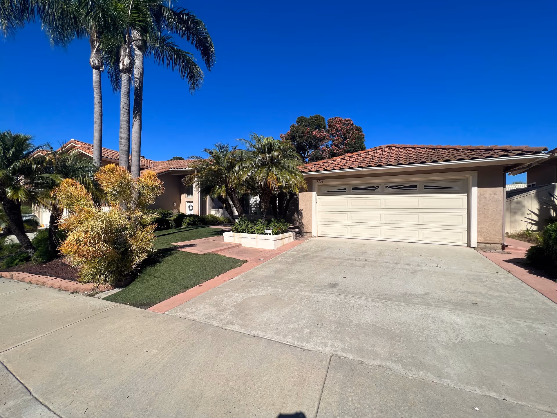 Front exterior view of a single-story house with a tiled roof, a two-car garage, and a driveway. The yard features palm trees, various shrubs, and a well-maintained lawn under a clear blue sky.
