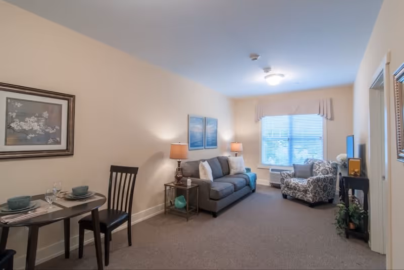 A cozy living room area with a gray sofa, two side tables with lamps, a patterned armchair near a window with blinds, a small dining table set for two with dishes and glasses, and framed artwork on the walls.