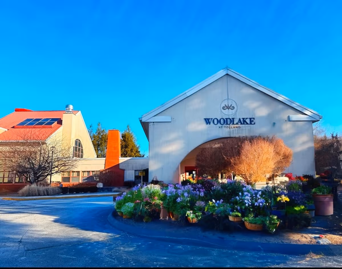 Front entrance of the Woodlake at Tolland nursing and rehabilitation center with a gabled entryway and a large display of potted flowers in front.