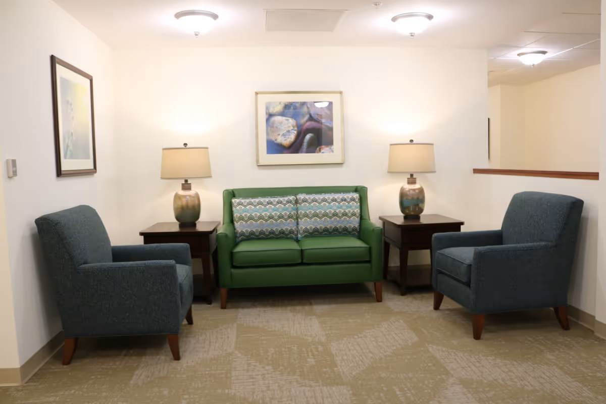 A small sitting area with a green loveseat adorned with two patterned cushions, flanked by two dark wooden side tables each holding a ceramic table lamp. Two blue upholstered armchairs face the loveseat. The walls are white with framed artwork, and the floor is covered with a beige patterned carpet.