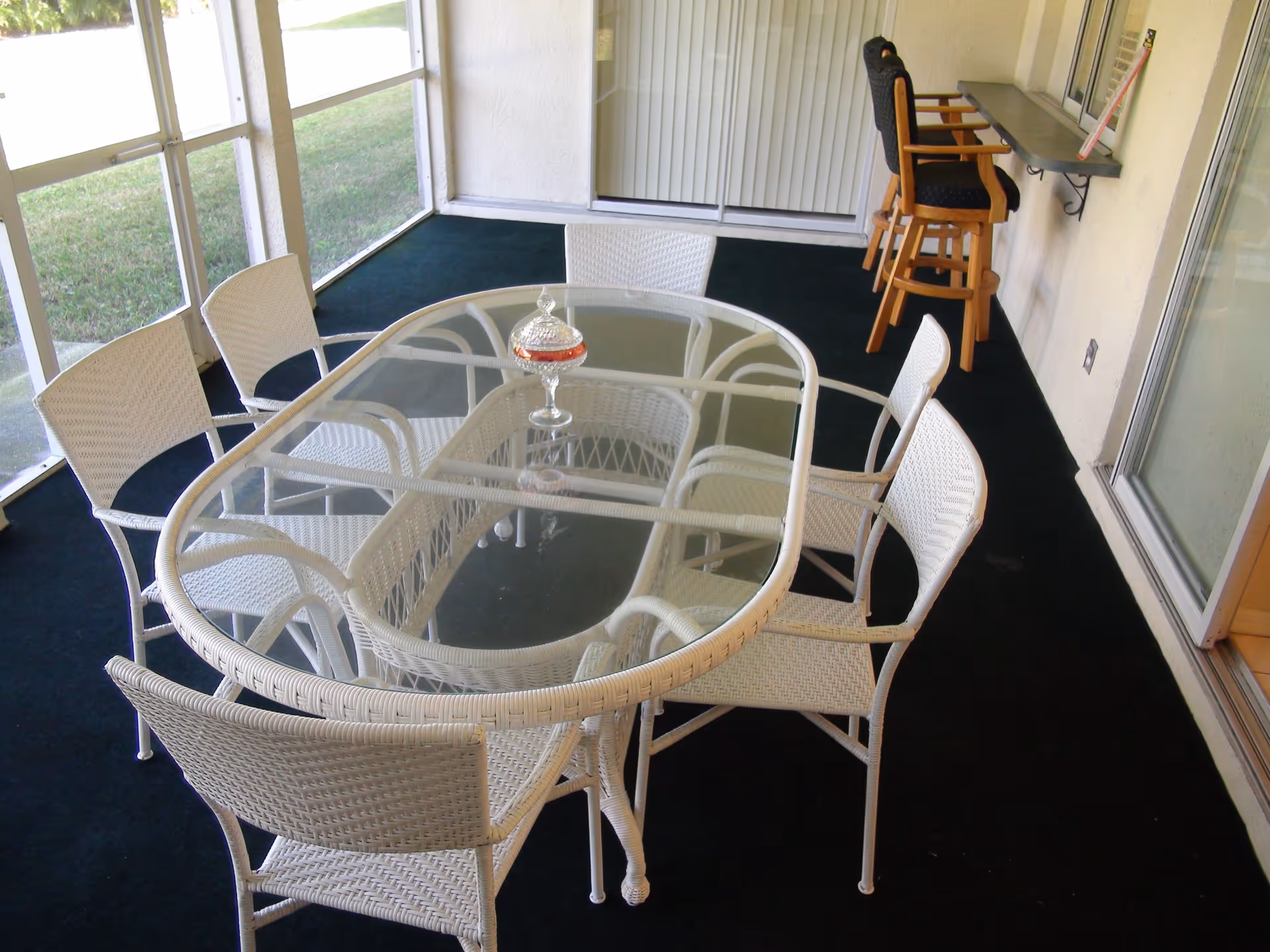 A screened-in porch area with a glass-top wicker dining table surrounded by six white wicker chairs. There are two wooden bar stools with black cushions near a small counter attached to the wall. The floor is covered with dark carpeting and there is a grassy lawn visible outside the screened windows.