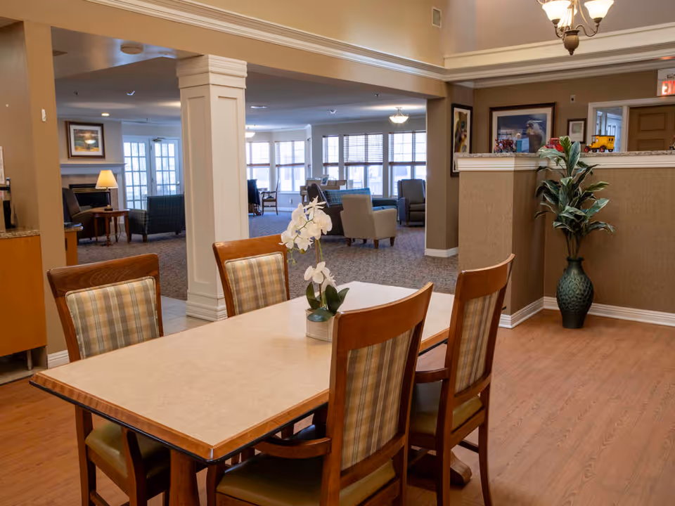 A well-lit interior space featuring a rectangular dining table with four wooden chairs upholstered in plaid fabric. A white orchid plant in a small pot is placed on the table. In the background, there is a spacious living area with multiple armchairs, large windows with blinds, framed artwork on the walls, and a large green vase with a plant near a half wall. The flooring is a combination of wood and carpet, and the ceiling has a chandelier light fixture.