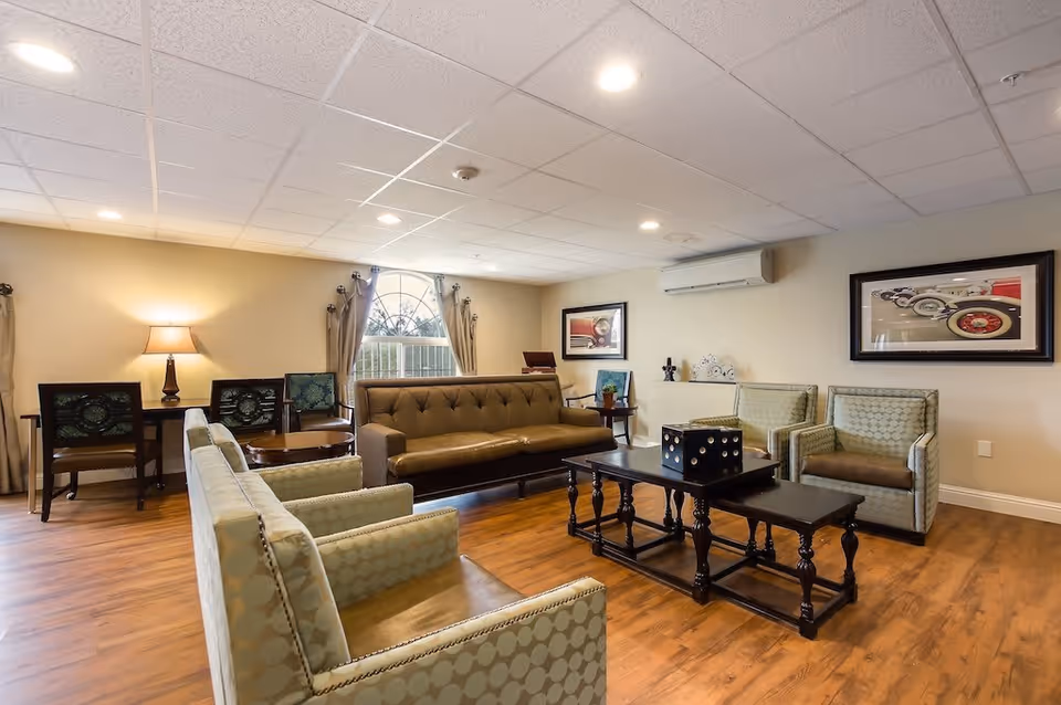 A cozy senior living facility common area with a brown leather sofa, patterned armchairs, a wooden coffee table with a large decorative dice, two framed pictures on the walls, a window with curtains, a desk with two chairs and a lamp, and wood flooring.