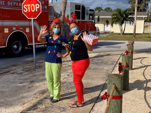 Two women wearing face masks and festive holiday accessories, including reindeer antlers and light bulb necklaces, stand on a sidewalk waving. Behind them is a red fire truck and a stop sign, with a residential house and palm trees in the background.