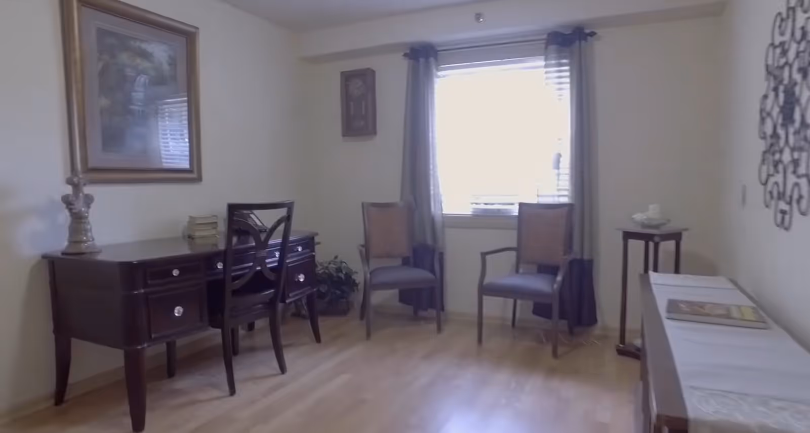 A small room with light-colored walls and wooden flooring featuring a dark wooden desk with a chair, two armchairs placed near a window with curtains, a small side table with a decorative item, a wall clock, a framed painting, and a decorative wall piece.