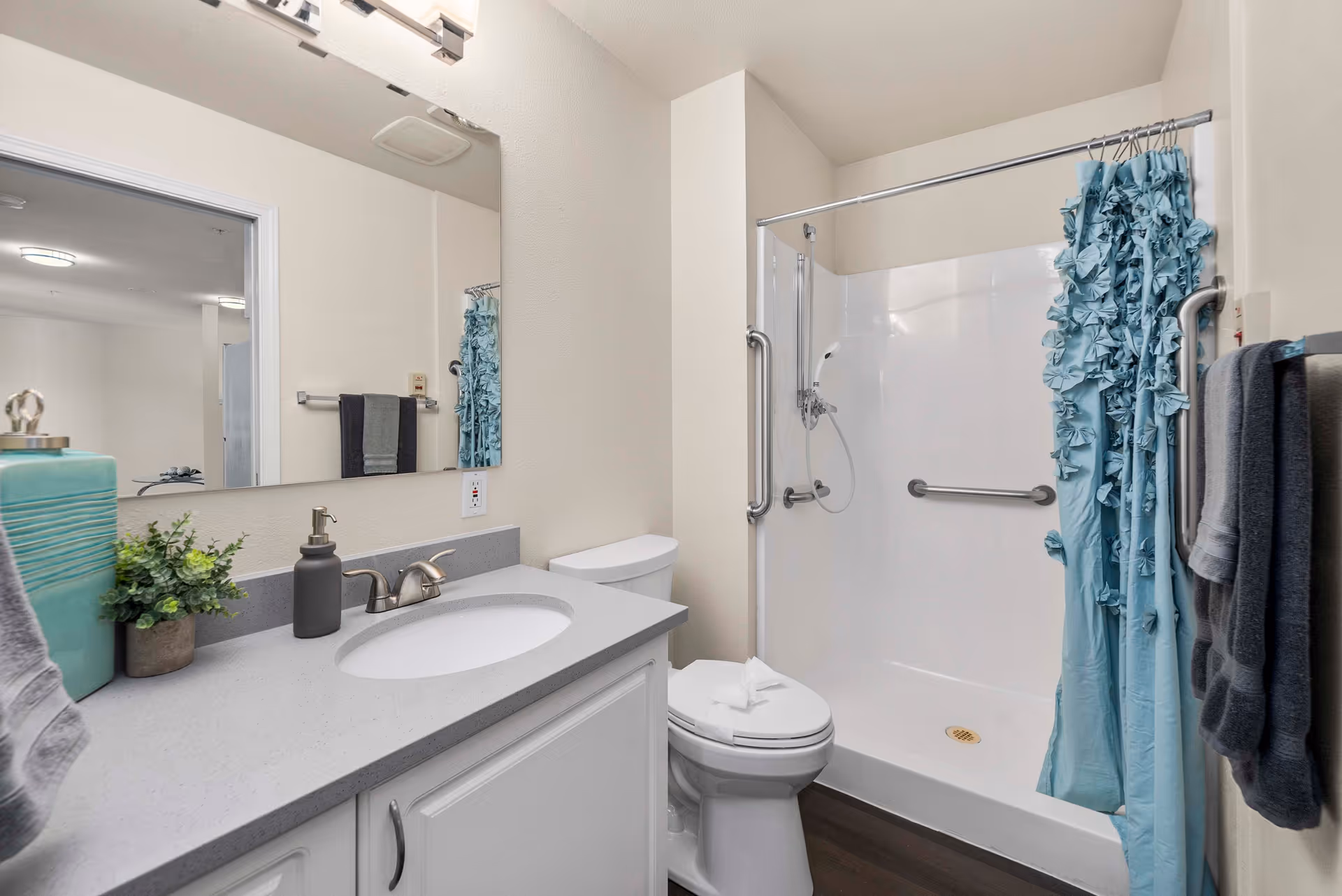 Accessible bathroom with a sink vanity, toilet, and a shower with a blue ruffled curtain and grab bars.