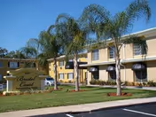 Exterior view of a two-story senior living facility building with beige walls and black awnings over the windows. There are several palm trees and a well-maintained lawn in front of the building. A sign near the entrance reads 'Bristol Court Senior Living'.