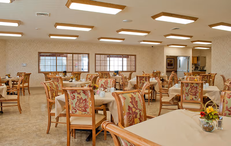 A spacious dining room with multiple tables covered in beige tablecloths, each surrounded by wooden chairs with floral upholstery. The room has patterned wallpaper, large windows, and ceiling lights. Small flower arrangements and condiments are placed on the tables.