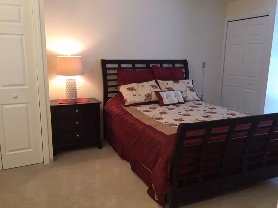 A bedroom with a dark wooden bed frame and a bed made with a floral and burgundy comforter set. There is a matching nightstand with a lamp on it to the left of the bed. The room has beige carpet and white walls, with two closed white closet doors visible.