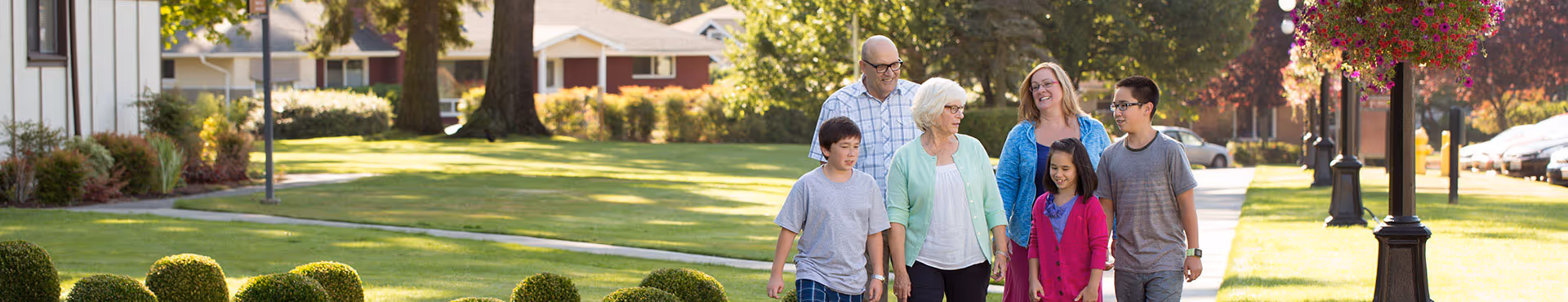 A group of six people, including older adults and children, walking together on a sidewalk in a green, sunny outdoor area with trees, bushes, and houses in the background.