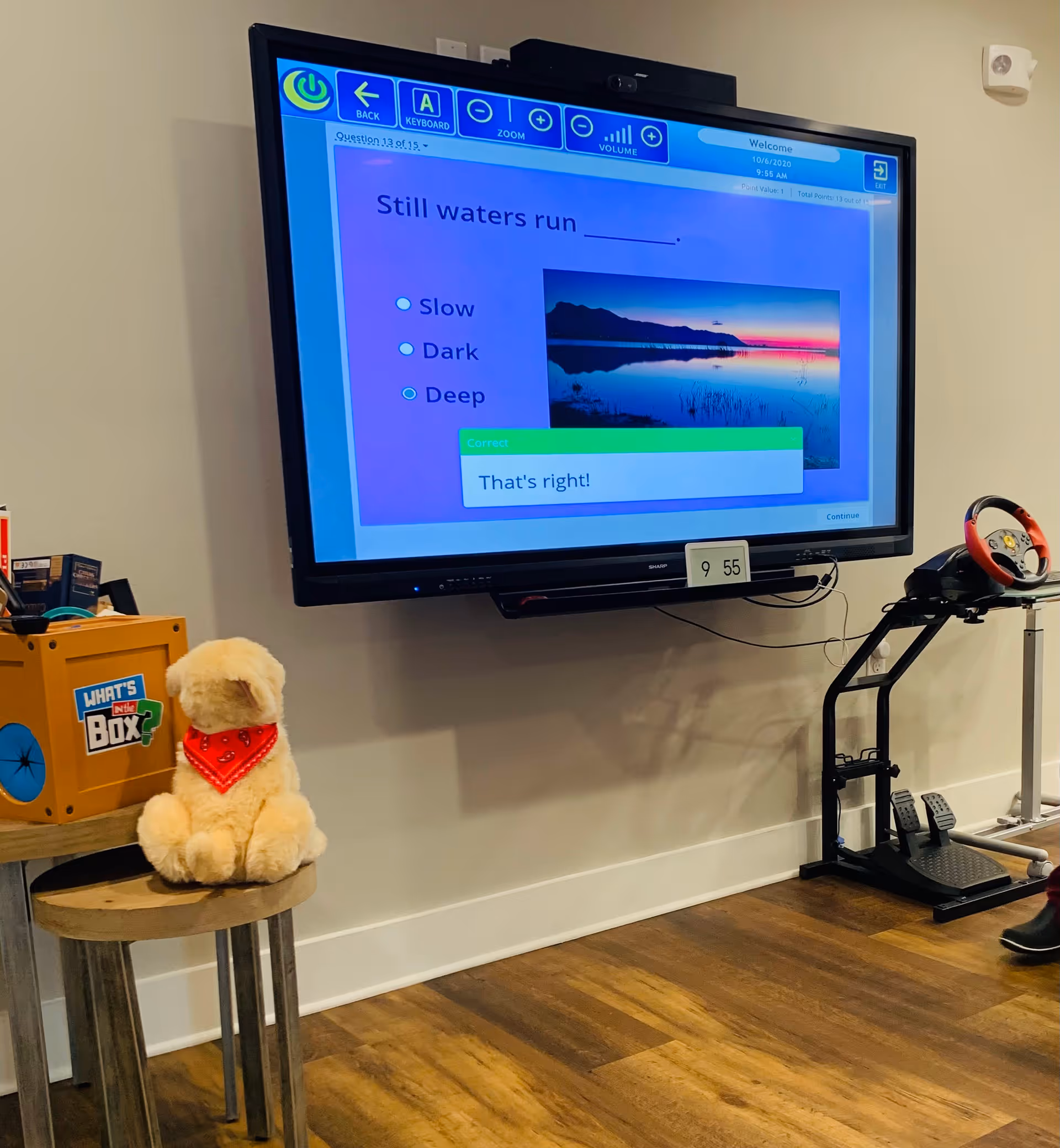 A wall-mounted large screen displaying a quiz question 'Still waters run ______.' with answer options Slow, Dark, Deep, and a confirmation message 'That's right!'. Below the screen is a digital clock showing 9:55. To the left, a small wooden table holds a plush dog toy wearing a red bandana and a box labeled 'What's in the Box?'. To the right, there is a gaming steering wheel setup with pedals on the floor.