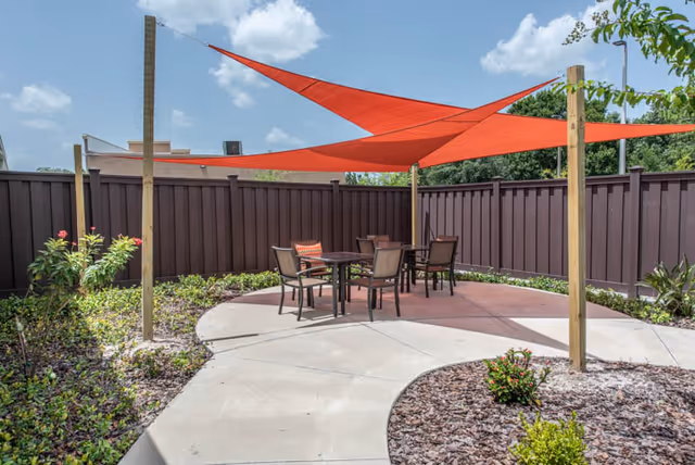 Outdoor patio area with a round concrete seating space featuring a table and four chairs under two large orange triangular shade sails. The area is surrounded by a brown privacy fence, landscaping with small bushes and plants, and a partly cloudy blue sky overhead.
