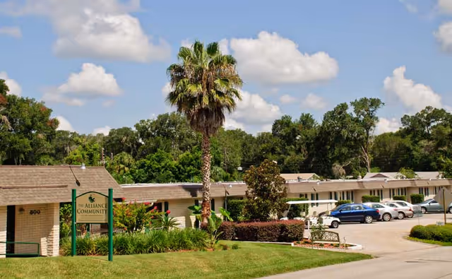 Exterior view of The Alliance Community senior living facility showing a single-story building with a row of parked cars, a palm tree, green bushes, and a sign with the facility's name under a partly cloudy sky.