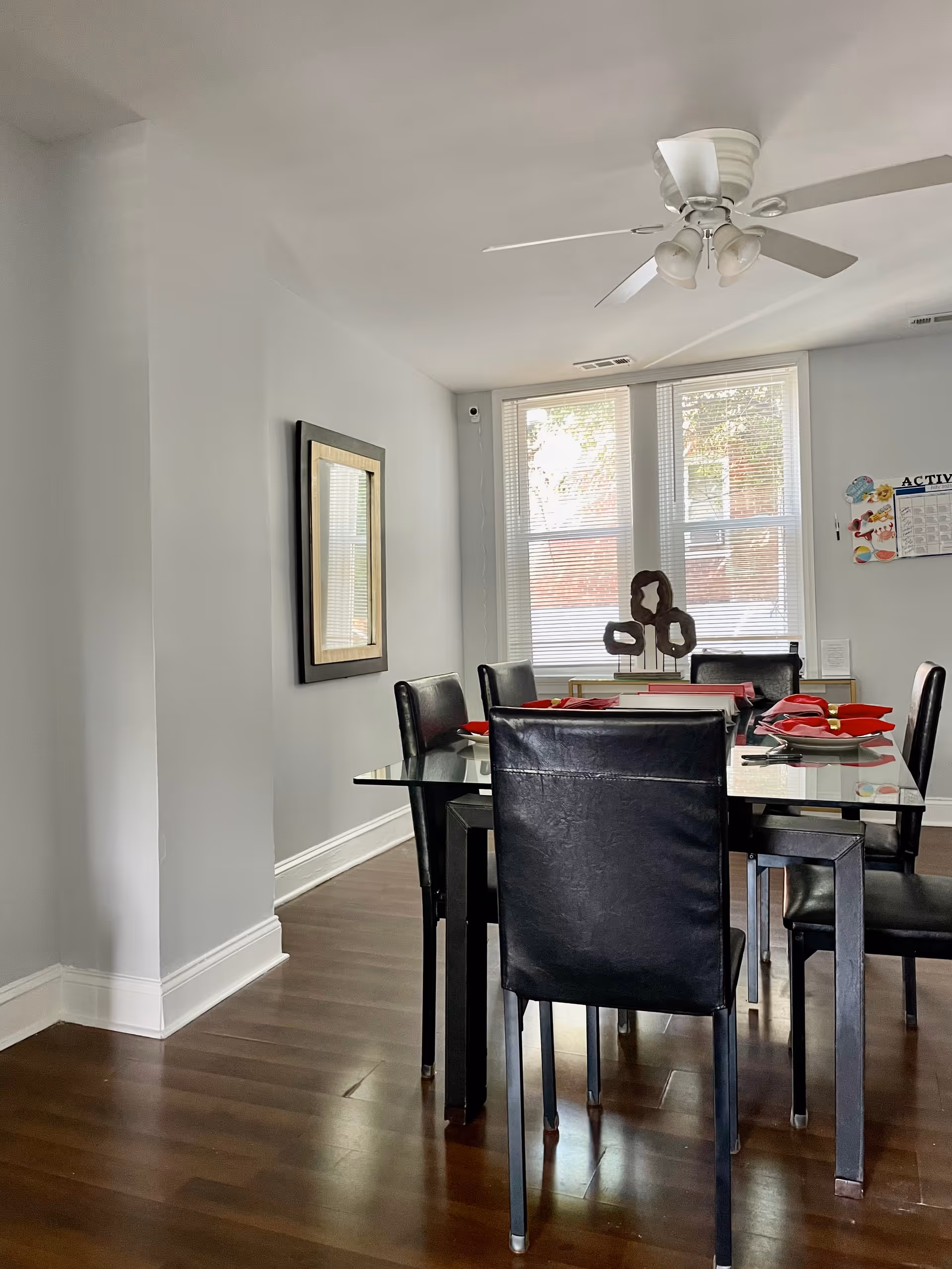 A dining room with a glass table set for six people with black leather chairs. The table has red napkins and a decorative centerpiece. There is a ceiling fan above, a framed mirror on the left wall, and three large windows with blinds letting in natural light.