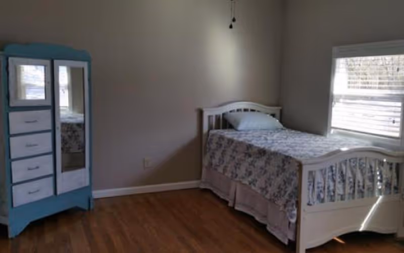 A simple bedroom with a single white bed covered with a floral bedspread and a pillow, positioned near a window with blinds. To the left of the bed is a blue dresser with drawers and a mirror. The room has wooden flooring and plain light-colored walls.
