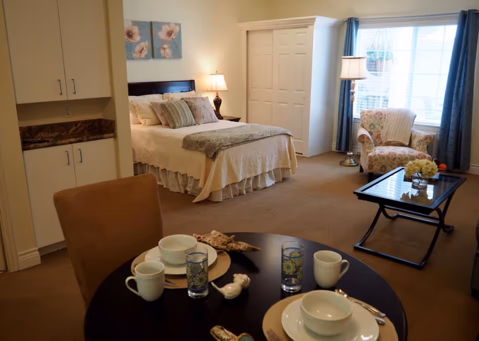 Furnished studio apartment interior with a bed, dining table set, armchair and coffee table by a large window.