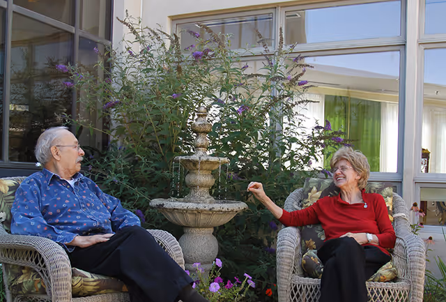 An elderly man and woman sitting on wicker chairs outside near a stone water fountain surrounded by plants and flowers, with a building window in the background. The woman is smiling and gesturing towards the man.