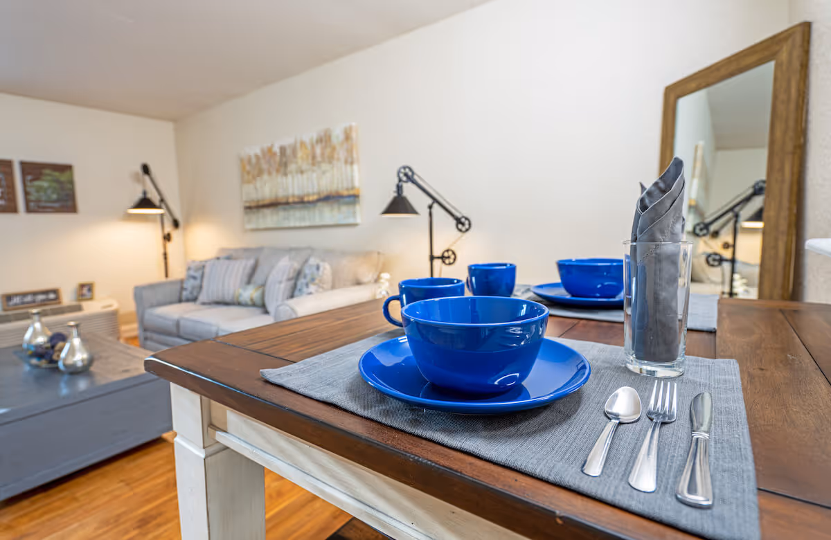 A close-up view of a wooden dining table set with blue bowls, plates, mugs, and silverware on gray placemats. In the background, there is a cozy living room with a gray sofa, decorative pillows, a floor lamp, a coffee table with decorative items, and a large mirror on the wall.