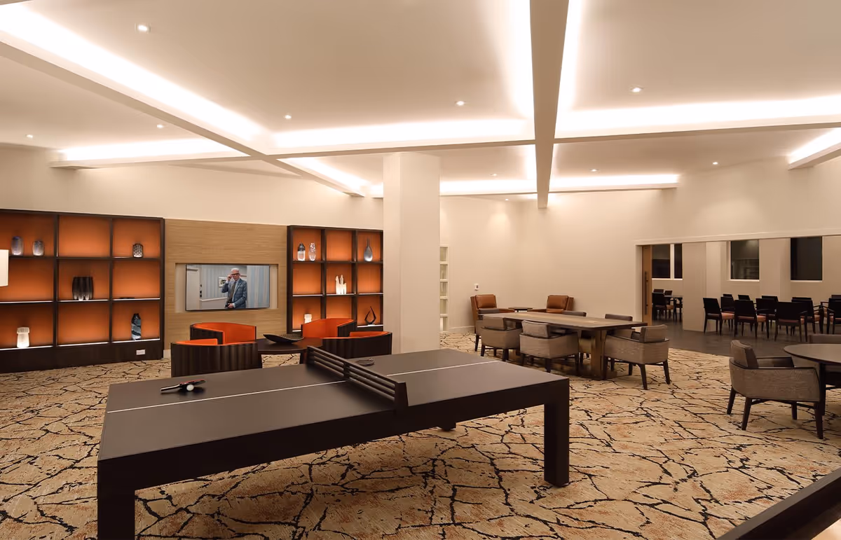 Spacious modern community lounge with a ping-pong table, seating areas, display shelving and warm recessed lighting.