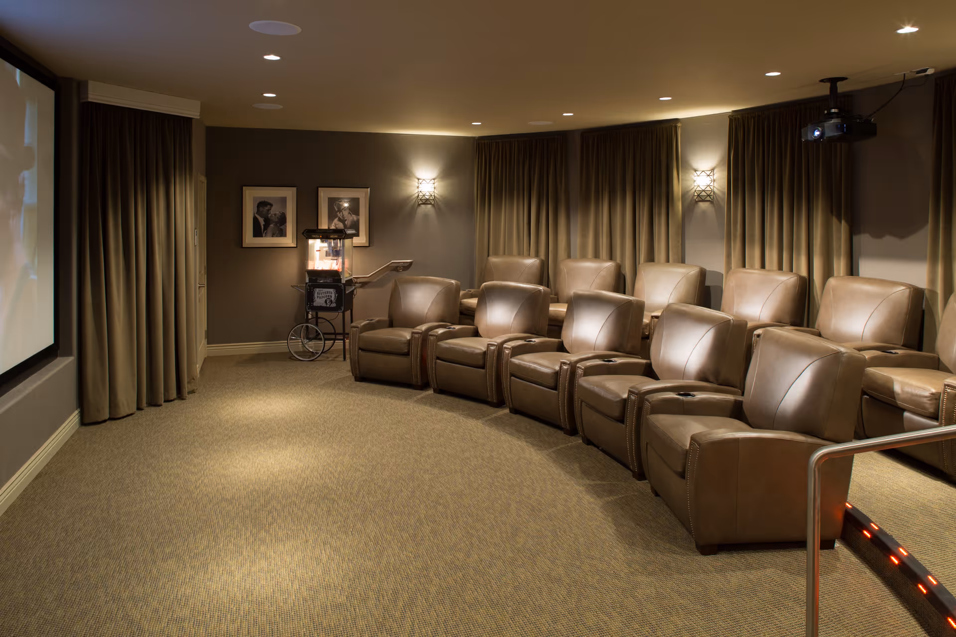 A cozy home theater room with two rows of brown leather recliner chairs facing a large projection screen. The room has beige carpet, dark curtains covering the windows, wall sconces providing soft lighting, and a popcorn machine in the back corner.
