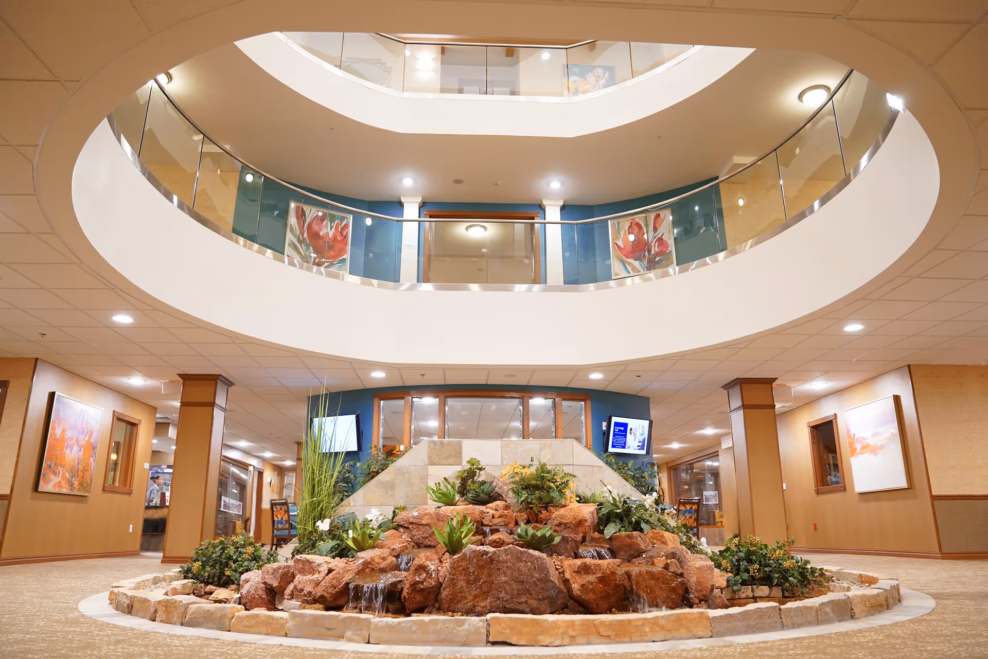 Interior view of a senior living facility featuring a circular atrium with a glass railing on the upper floor. In the center of the ground floor is a decorative rock water feature with small waterfalls and green plants. The surrounding area has beige walls, carpeted floors, and framed artwork on the walls.